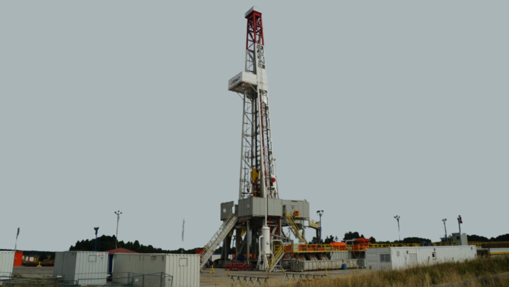 A large oil drilling rig situated in a field, surrounded by equipment containers and safety barriers under an overcast sky.